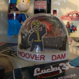 May include: A clear snow globe featuring a cartoon character in a yellow hard hat, blue suit, and yellow gloves, with the text "HOOVER DAM" in red. The background shows a dam structure.
