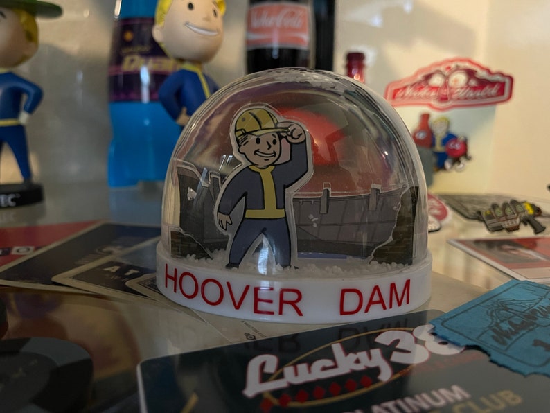 May include: A clear snow globe featuring a cartoon character in a yellow hard hat, blue suit, and yellow gloves, with the text "HOOVER DAM" in red. The background shows a dam structure.