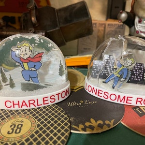 May include: Two snow globes with white bases and clear domes. One features a cartoon character in a blue jumpsuit and red scarf with the text "MT. CHARLESTON". The other shows the same character with the text "LONESOME ROAD".