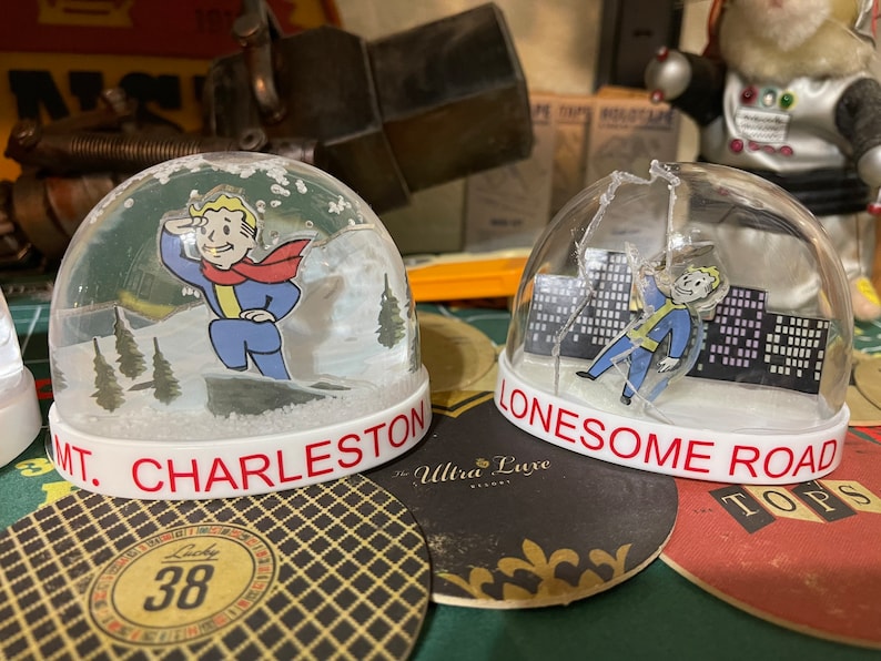 May include: Two snow globes with white bases and clear domes. One features a cartoon character in a blue jumpsuit and red scarf with the text "MT. CHARLESTON". The other shows the same character with the text "LONESOME ROAD".