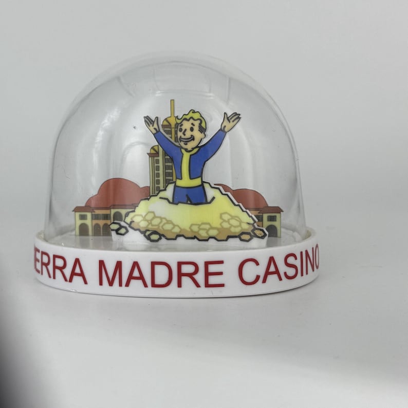 May include: A clear, dome-shaped snow globe with a white base. The base reads "ERRA MADRE CASINO" in red letters. Inside, a cartoon figure in a blue jumpsuit is depicted with arms raised. The background features a building and a cloud.