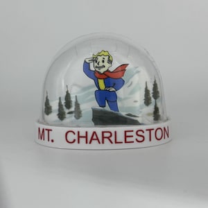 May include: A snow globe featuring a cartoon character in a blue jumpsuit and red scarf, saluting against a snowy mountain backdrop. The base is white with the text "MT. CHARLESTON" in red.