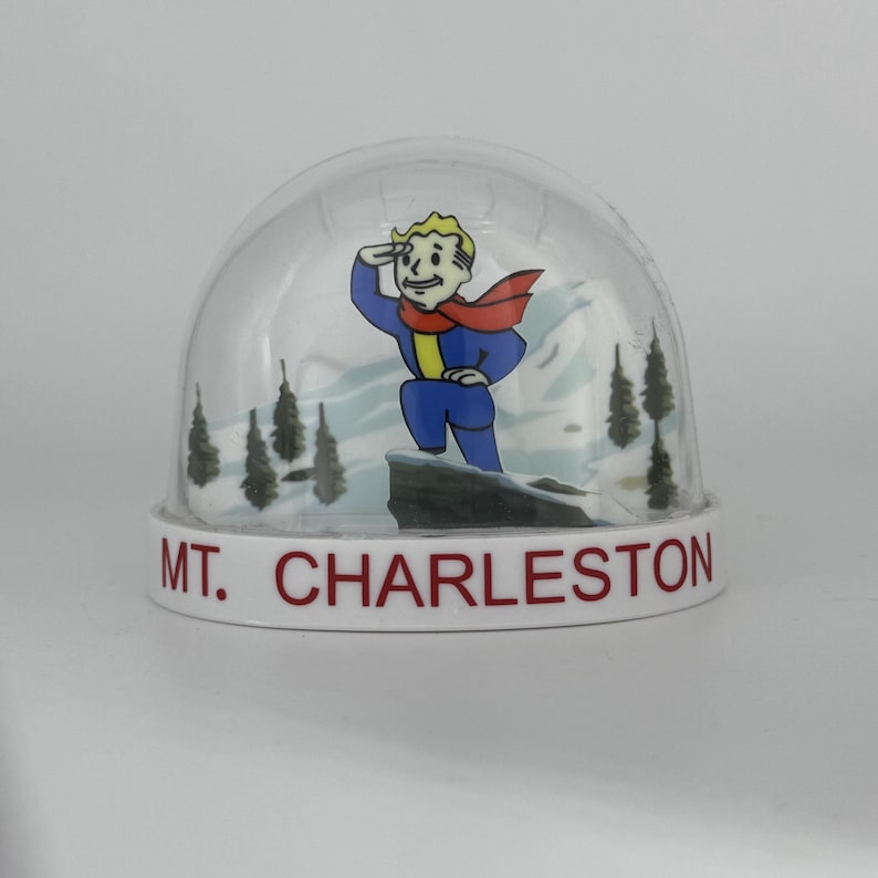 May include: A snow globe featuring a cartoon character in a blue jumpsuit and red scarf, saluting against a snowy mountain backdrop. The base is white with the text "MT. CHARLESTON" in red.