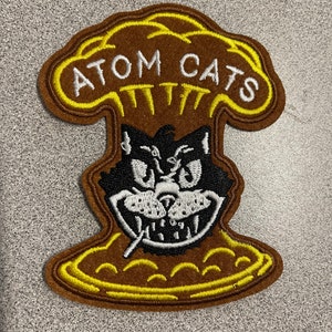 Peut inclure: Un écusson brodé marron et jaune avec le texte "ATOM CATS" en lettres blanches. L'écusson représente un chat de dessin animé avec un sourire malicieux et une cigarette à la bouche.