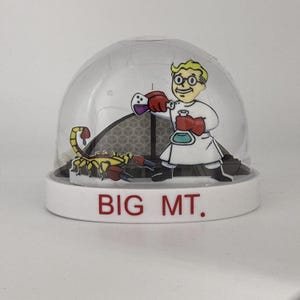 May include: A snow globe with a white base and a clear dome. Inside, a cartoon scientist in a lab coat and a scorpion are featured. The base reads "BIG MT." in red letters.