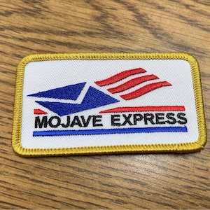 Op de afbeelding: Een witte geborduurde patch met een gestileerd blauw en rood envelopontwerp en de tekst "MOJAVE EXPRESS" in zwarte letters. De patch heeft een gouden rand.