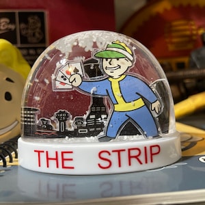 May include: A clear snow globe featuring a cartoon character in a blue and yellow outfit holding playing cards. The base of the globe reads "THE STRIP" in red letters. The background includes a black and white cityscape and falling white flakes.