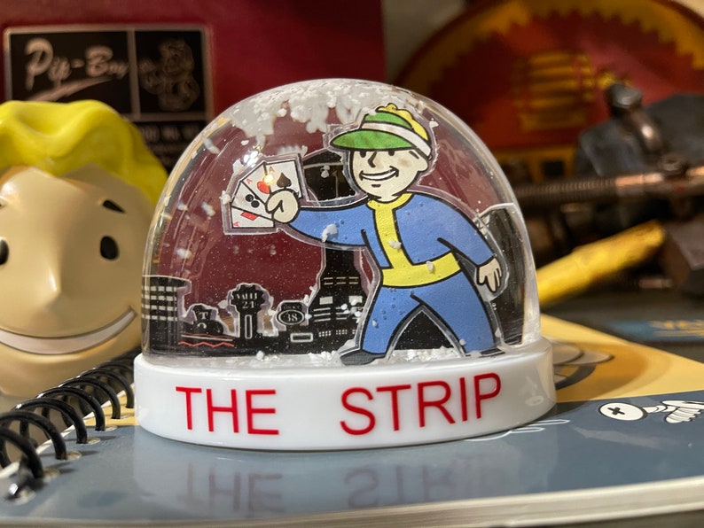 May include: A clear snow globe featuring a cartoon character in a blue and yellow outfit holding playing cards. The base of the globe reads "THE STRIP" in red letters. The background includes a black and white cityscape and falling white flakes.