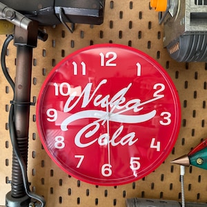 May include: A red, round wall clock with the words "Nuka Cola" in white script. The clock has white numbers and hands, and is mounted on a brown pegboard wall. The clock is a retro-style item.