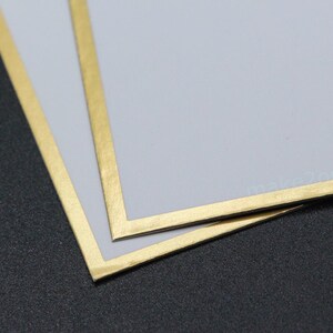 May include: Two pale grey cards with gold borders are stacked on a dark grey surface. The gold borders frame the edges of the cards. The cards slightly overlap, showing the gold border and the grey surface.