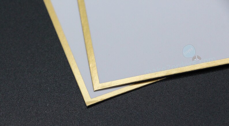 May include: Two pale grey cards with gold borders are stacked on a dark grey surface. The gold borders frame the edges of the cards. The cards slightly overlap, showing the gold border and the grey surface.
