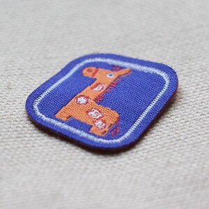 May include: A square patch with a cartoon giraffe design. The giraffe is orange with white spots, set on a blue background with a white border. Suitable for clothing or accessories.