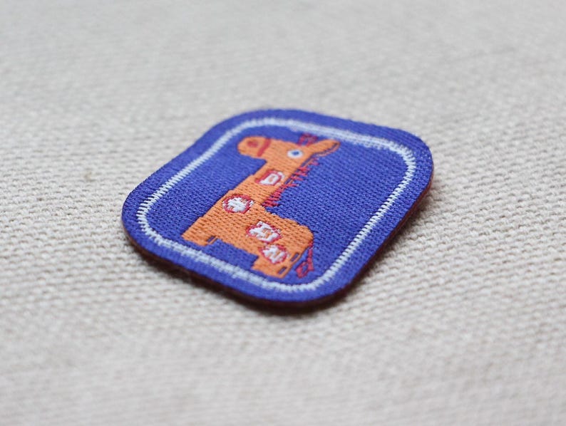 May include: A square patch with a cartoon giraffe design. The giraffe is orange with white spots, set on a blue background with a white border. Suitable for clothing or accessories.