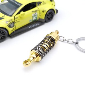 May include: A gold and black metal keychain shaped like a car suspension component. The keychain is attached to a silver metal ring. A yellow toy race car is in the background.