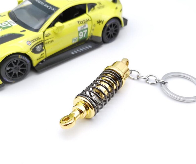 May include: A gold and black metal keychain shaped like a car suspension component. The keychain is attached to a silver metal ring. A yellow toy race car is in the background.