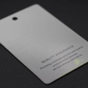 May include: A rectangular, light gray tag with a hole at the top. The text "QUALITY ASSURANCE" is printed on the tag, along with a statement about providing quality products. The tag is on a dark surface.