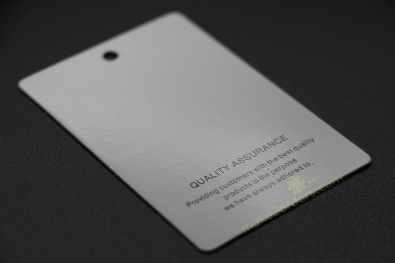 May include: A rectangular, light gray tag with a hole at the top. The text "QUALITY ASSURANCE" is printed on the tag, along with a statement about providing quality products. The tag is on a dark surface.