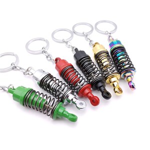 May include: A collection of six keychains featuring miniature shock absorber designs. The keychains showcase a variety of colors, including green, silver, red, black, gold, and iridescent. Each keychain has a silver chain and ring for attachment.