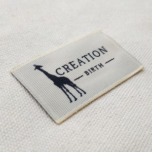 May include: A rectangular label with the words "CREATION" and "BIRTH" printed in navy blue. A navy blue giraffe silhouette is on the left side of the label. The label is on a white textured surface.