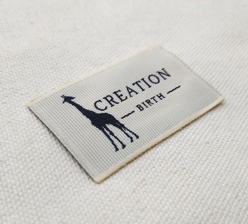 May include: A rectangular label with the words "CREATION" and "BIRTH" printed in navy blue. A navy blue giraffe silhouette is on the left side of the label. The label is on a white textured surface.
