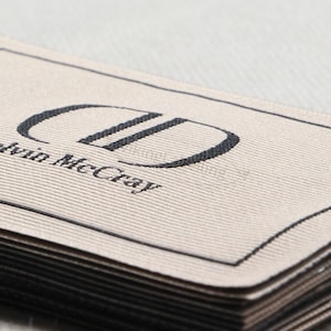 May include: Close-up of a stack of woven fabric labels. The top label is beige with a black border and the text "Delvin McCray" below a stylized "DD" logo. The labels are neatly stacked.