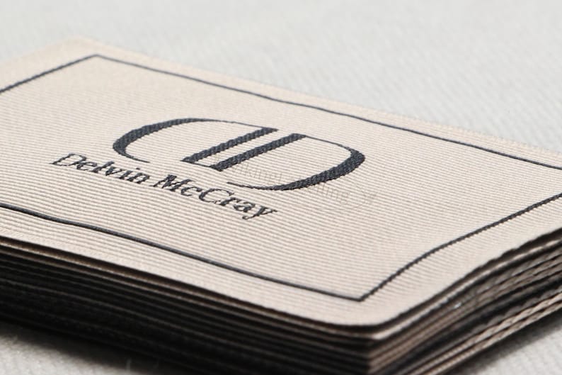 May include: Close-up of a stack of woven fabric labels. The top label is beige with a black border and the text "Delvin McCray" below a stylized "DD" logo. The labels are neatly stacked.
