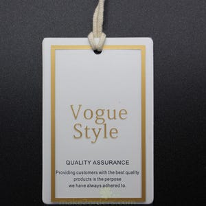 May include: A white rectangular tag with a gold border and the words "Vogue Style" in gold lettering. The tag includes the text "QUALITY ASSURANCE" and a statement about providing quality products. A cream-colored string is attached.
