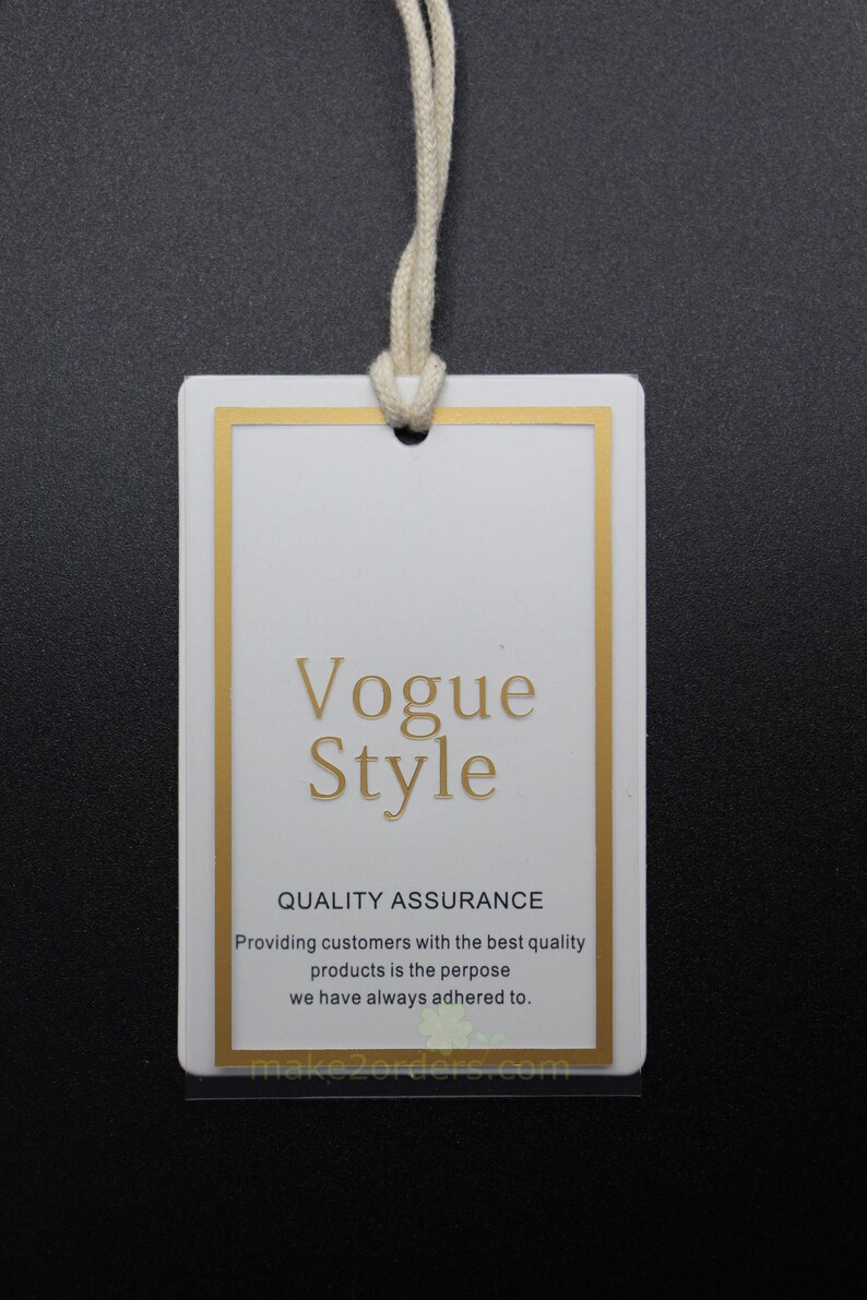 May include: A white rectangular tag with a gold border and the words "Vogue Style" in gold lettering. The tag includes the text "QUALITY ASSURANCE" and a statement about providing quality products. A cream-colored string is attached.