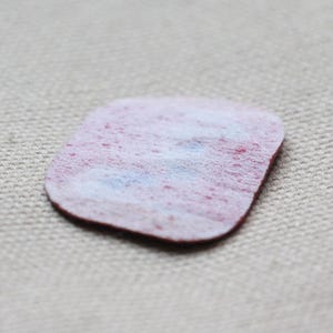 May include: A small, irregularly shaped, felt patch with a red border and a white and red mottled centre. The patch is set against a neutral, textured background.