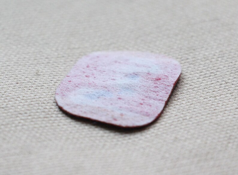 May include: A small, irregularly shaped, felt patch with a red border and a white and red mottled centre. The patch is set against a neutral, textured background.