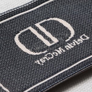 May include: Close-up of a rectangular label with a dark blue woven texture. The label features a white border and a white embroidered logo with the text "Dior" and "Dior Maison" in a stylized font.