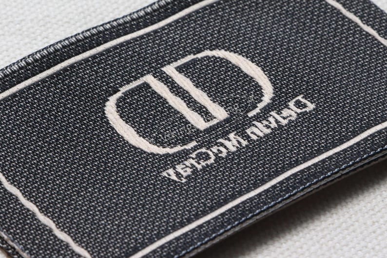 May include: Close-up of a rectangular label with a dark blue woven texture. The label features a white border and a white embroidered logo with the text "Dior" and "Dior Maison" in a stylized font.