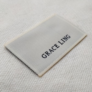 May include: A rectangular woven label with the text "GRACE LING" in black. The label has a light gray, ribbed texture and a cream-colored border. The label is placed on a textured, off-white fabric.