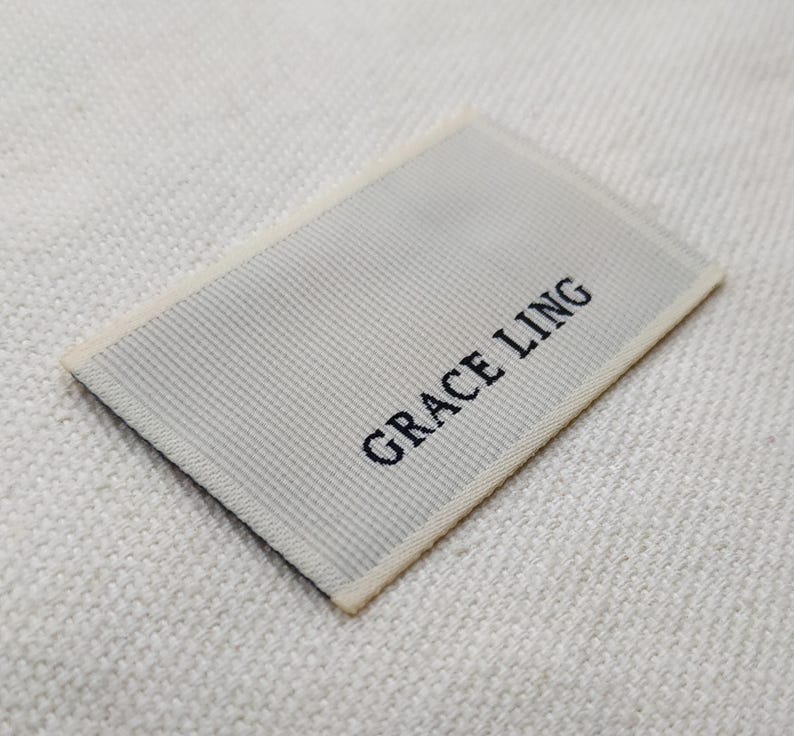 May include: A rectangular woven label with the text "GRACE LING" in black. The label has a light gray, ribbed texture and a cream-colored border. The label is placed on a textured, off-white fabric.