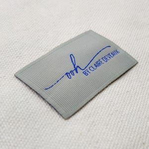 May include: A rectangular gray label with blue text that reads "ooh BY CLAIRE DEVEAUX". The label has a ribbed texture and is slightly curved. The text is in a handwritten style.
