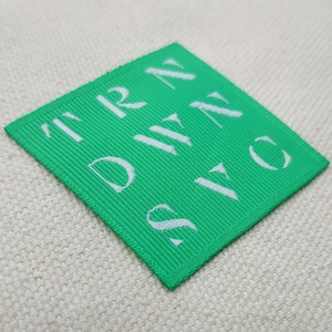 May include: A square, bright green label with white text reading "TRN DWN SVC". The label is made of a ribbed fabric and is set against a neutral, textured background.