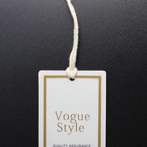 May include: A white rectangular clothing tag with gold trim and a cream-colored string. The tag features the words "Vogue Style" in gold lettering, along with additional text about quality assurance.
