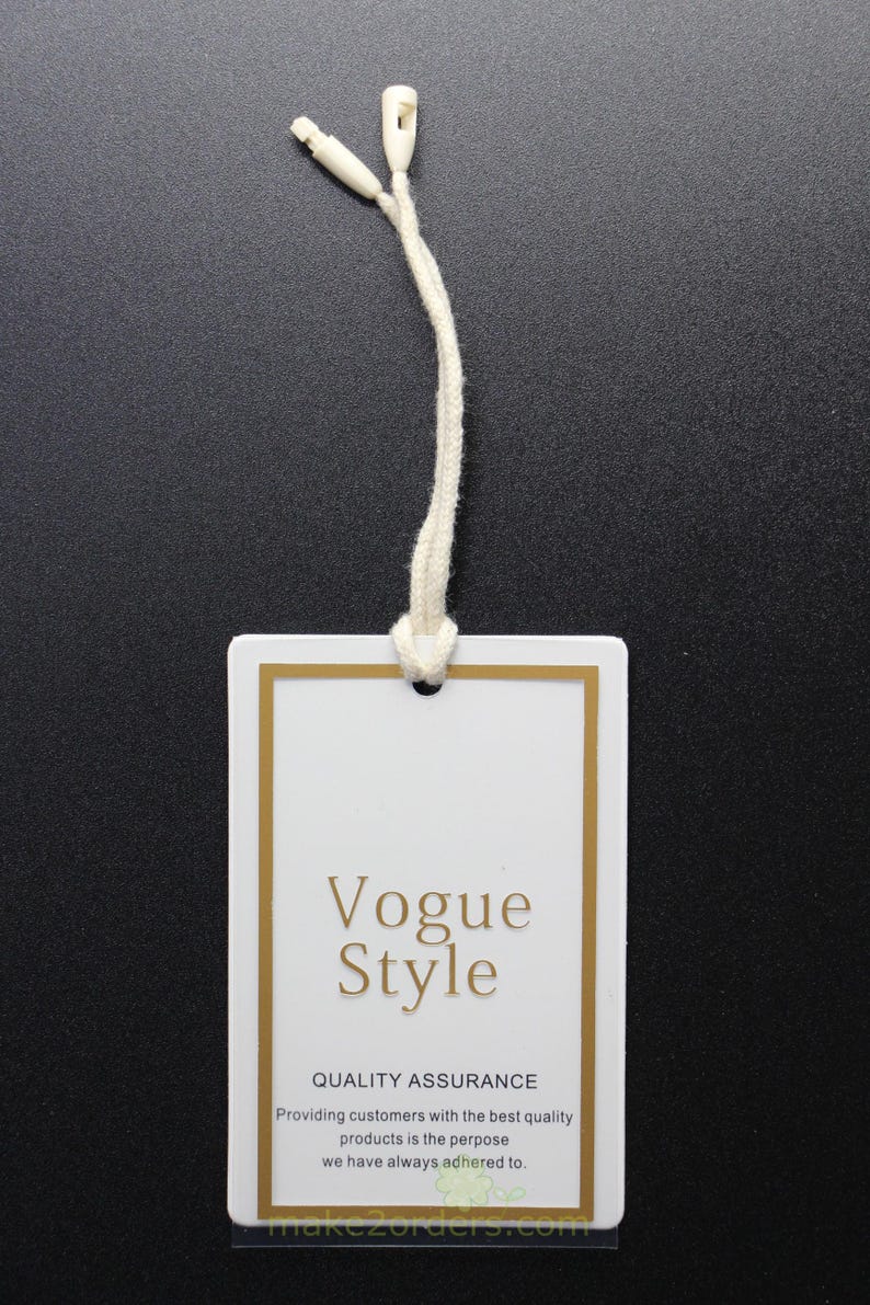 May include: A white rectangular clothing tag with gold trim and a cream-colored string. The tag features the words "Vogue Style" in gold lettering, along with additional text about quality assurance.