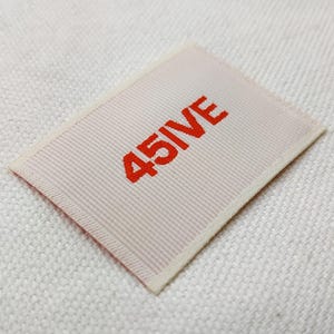 May include: A rectangular label with the text "45IVE" in bright red, centered on a light pink background. The label has a ribbed texture and a cream-colored border. The label is set against a white, textured fabric.