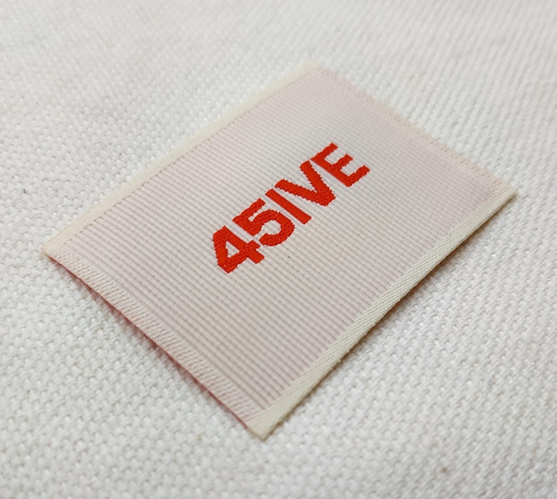 May include: A rectangular label with the text "45IVE" in bright red, centered on a light pink background. The label has a ribbed texture and a cream-colored border. The label is set against a white, textured fabric.