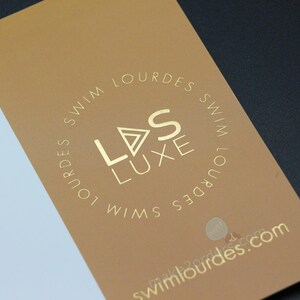 May include: A business card with a gold foil logo and text. The card is split into a white and a tan section. The logo reads "LDS LUXE SWIM LOURDES" in a circular pattern. The website address is also visible.