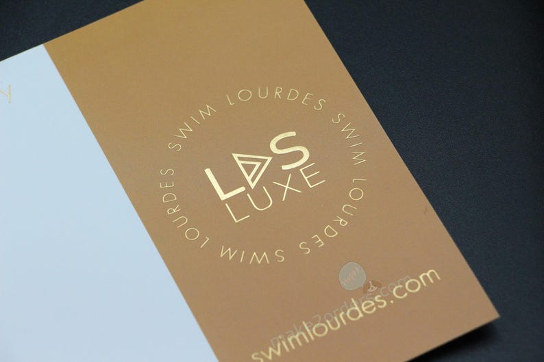 May include: A business card with a gold foil logo and text. The card is split into a white and a tan section. The logo reads "LDS LUXE SWIM LOURDES" in a circular pattern. The website address is also visible.