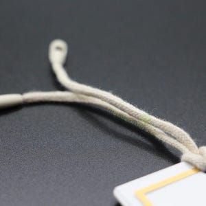 May include: Close-up of a beige drawstring cord with a plastic tip, attached to a white tag with a yellow border. The cord is looped and knotted, suggesting its use in clothing or accessories.
