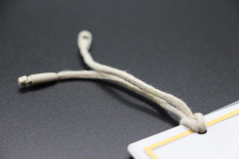 May include: Close-up of a beige drawstring cord with a plastic tip, attached to a white tag with a yellow border. The cord is looped and knotted, suggesting its use in clothing or accessories.