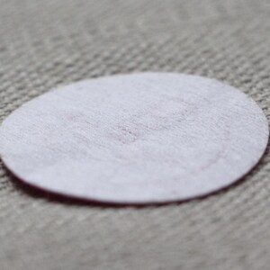 May include: A small, white, oval-shaped felt pad with a slightly pinkish hue in the centre. The pad is set against a textured, light grey fabric background. The felt pad is a simple, minimalist design.