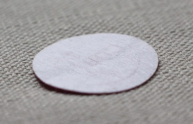 May include: A small, white, oval-shaped felt pad with a slightly pinkish hue in the centre. The pad is set against a textured, light grey fabric background. The felt pad is a simple, minimalist design.