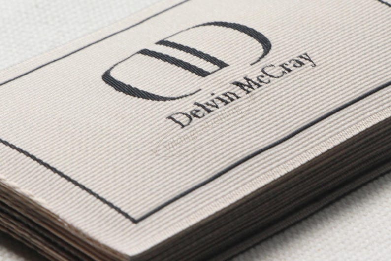 May include: Close-up of a stack of woven labels with the text "Delvin McCray" in a serif font. The logo features a stylized "D" in black. The labels have a light beige background with a black border.