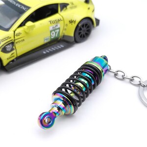 May include: A colorful keychain featuring a miniature shock absorber with a black spring and iridescent finish. The keychain is attached to a silver chain and ring. A yellow toy race car is in the background.