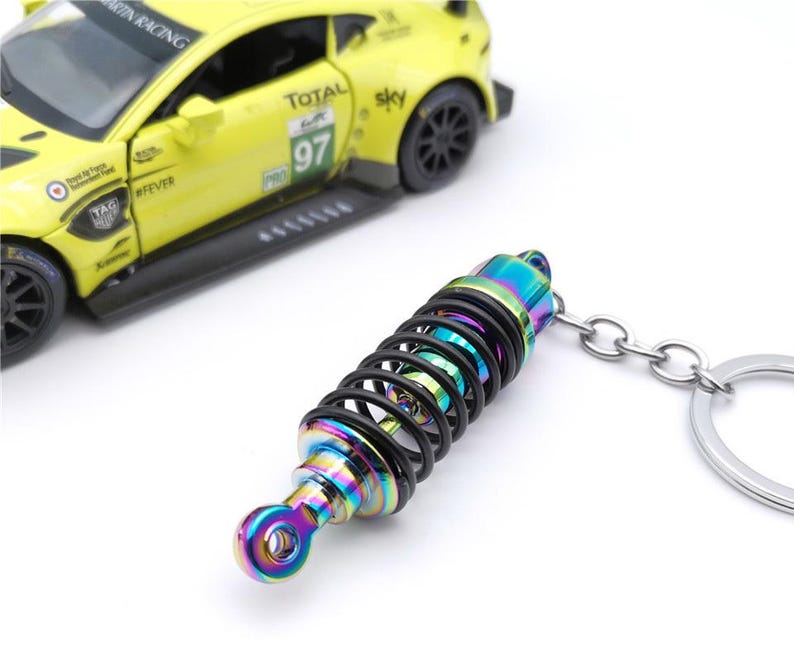 May include: A colorful keychain featuring a miniature shock absorber with a black spring and iridescent finish. The keychain is attached to a silver chain and ring. A yellow toy race car is in the background.