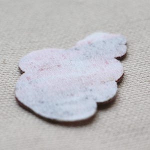 May include: A small, light-coloured felt appliqu&eacute; in a floral shape. The felt is white with a subtle pattern of pink and blue. The edges are a dark brown colour. The appliqu&eacute; is likely for crafting or embellishment.